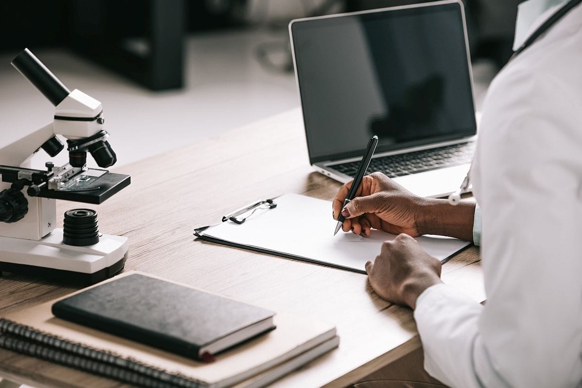 Health care professional seated at desk with laptop and microscope, writing; cost control concept