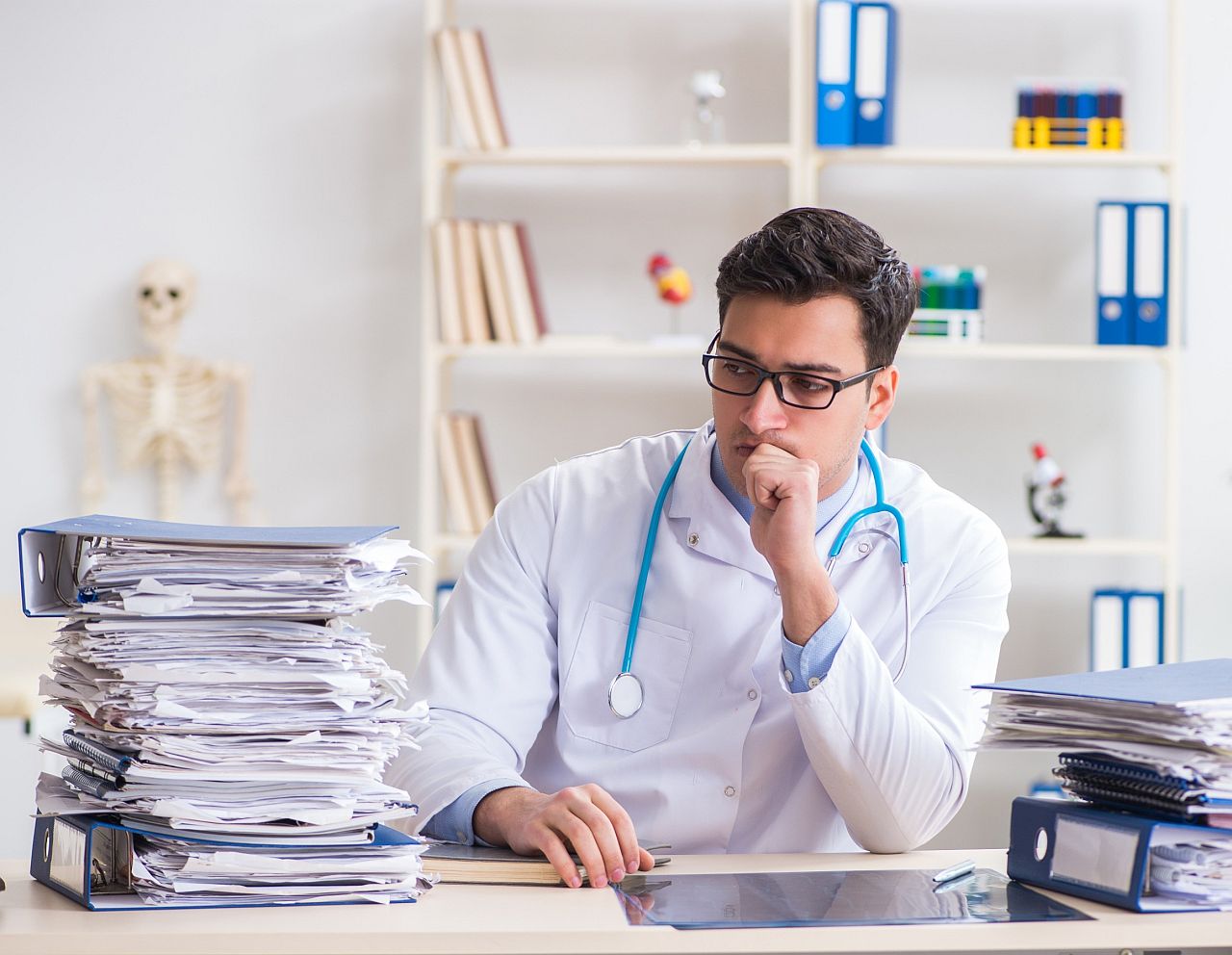Doctor with stethoscope around his neck, sitting at his desk, looks at stack of files and papers; specialty therapies concept