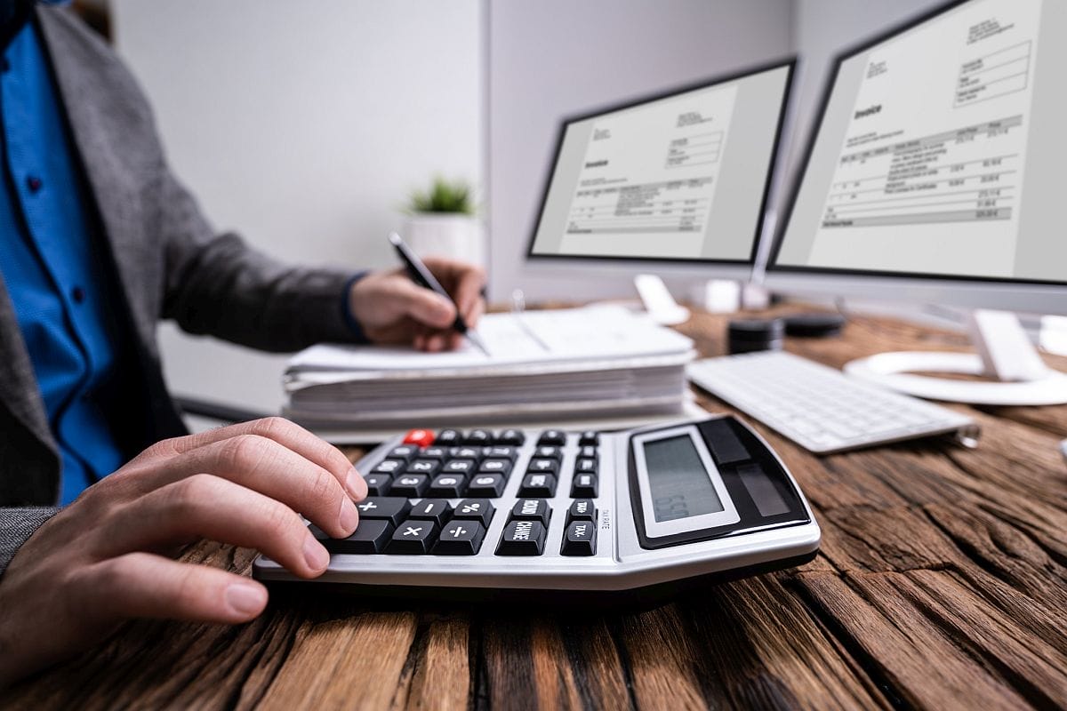 Man working on calculator with two monitors open and stack of papers; specialty therapies concept